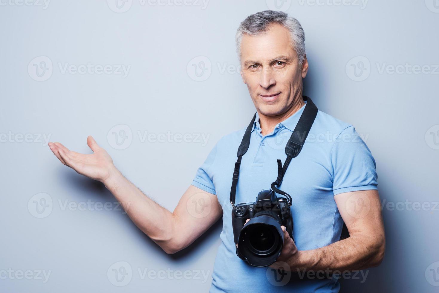 Here can be your advertising. Portrait of confident mature man in T-shirt holding camera and pointing away while standing against grey background photo