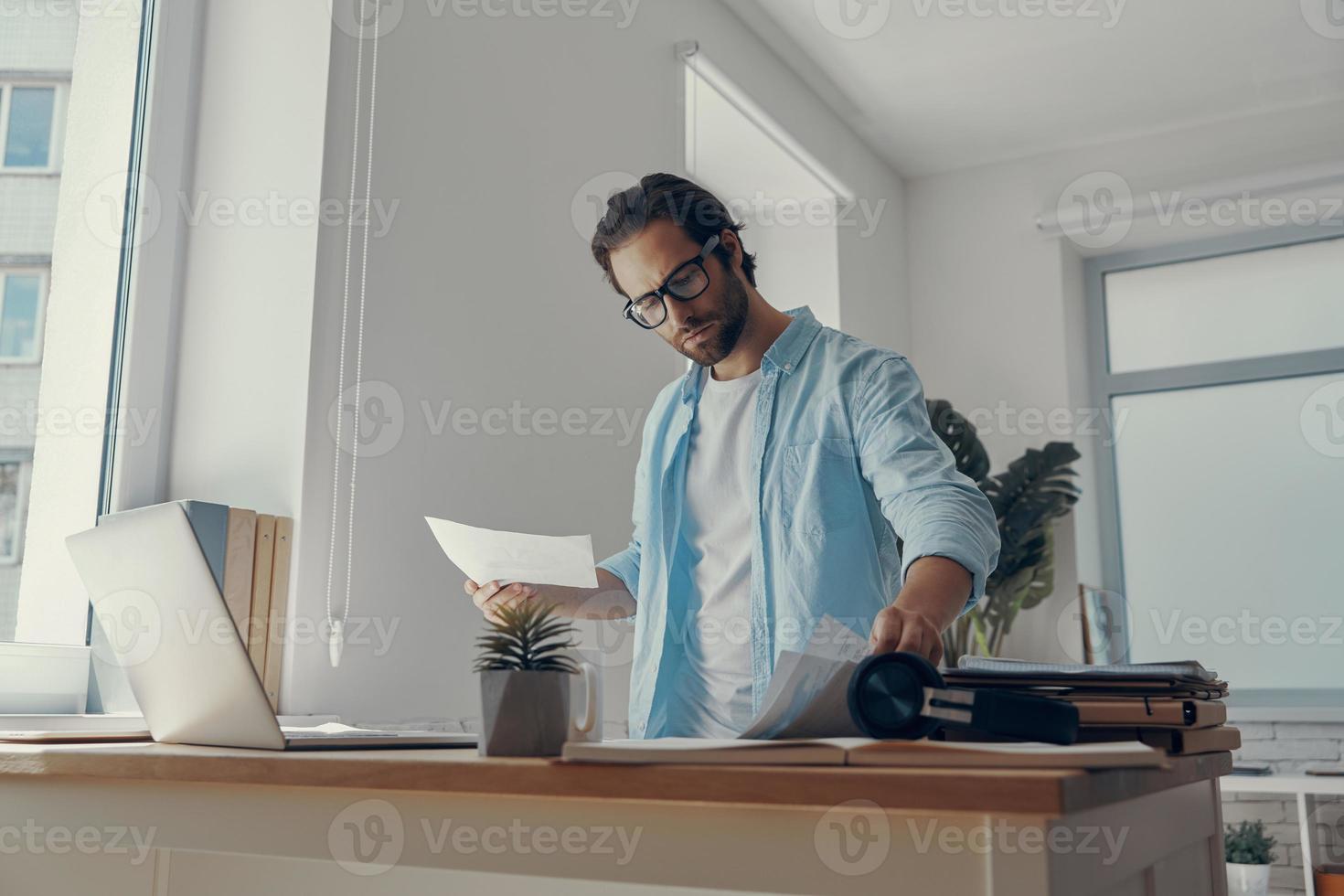 Confident young man doing some paperwork while standing near his working place in office photo