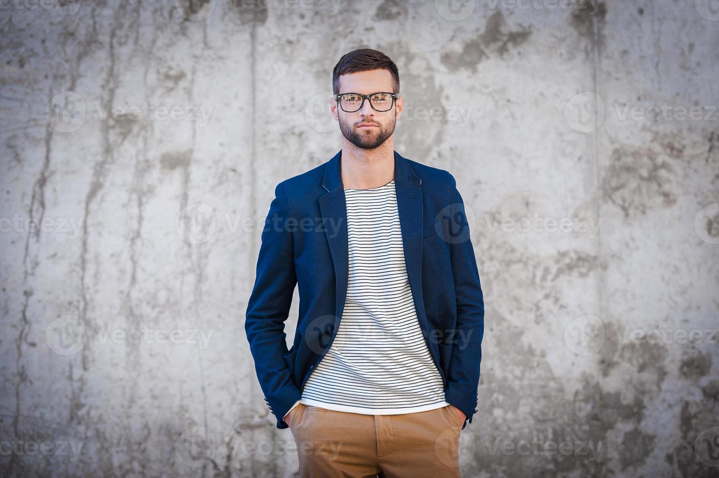 Feeling confident in my skin. Handsome young man in smart jacket holding hands in pockets while standing in front of the concrete wall photo