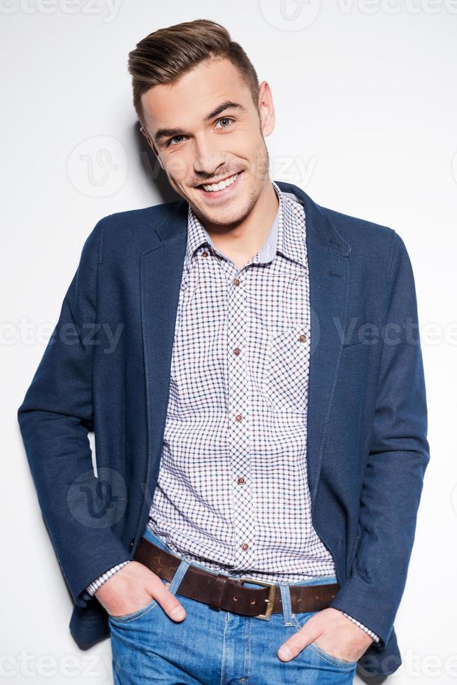Casually handsome. Handsome young man in smart casual wear holding hands in pockets and smiling while standing against a wall photo