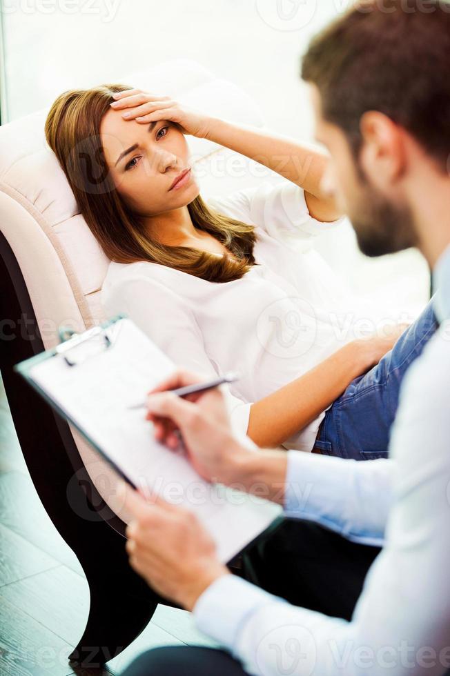 Feeling hopeless and depressed. Depressed young woman sitting at the chair and holding hand on head while young man sitting close to her and writing something in his clipboard photo