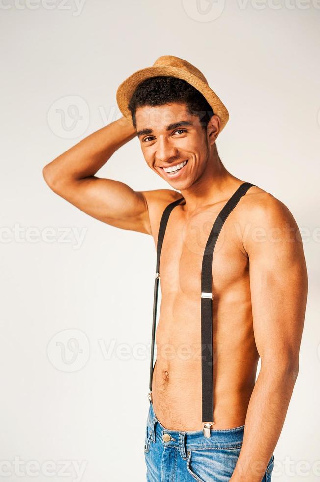 Young and free. Handsome young Afro-American hipster holding his hand on the hat and smiling while standing against grey background photo