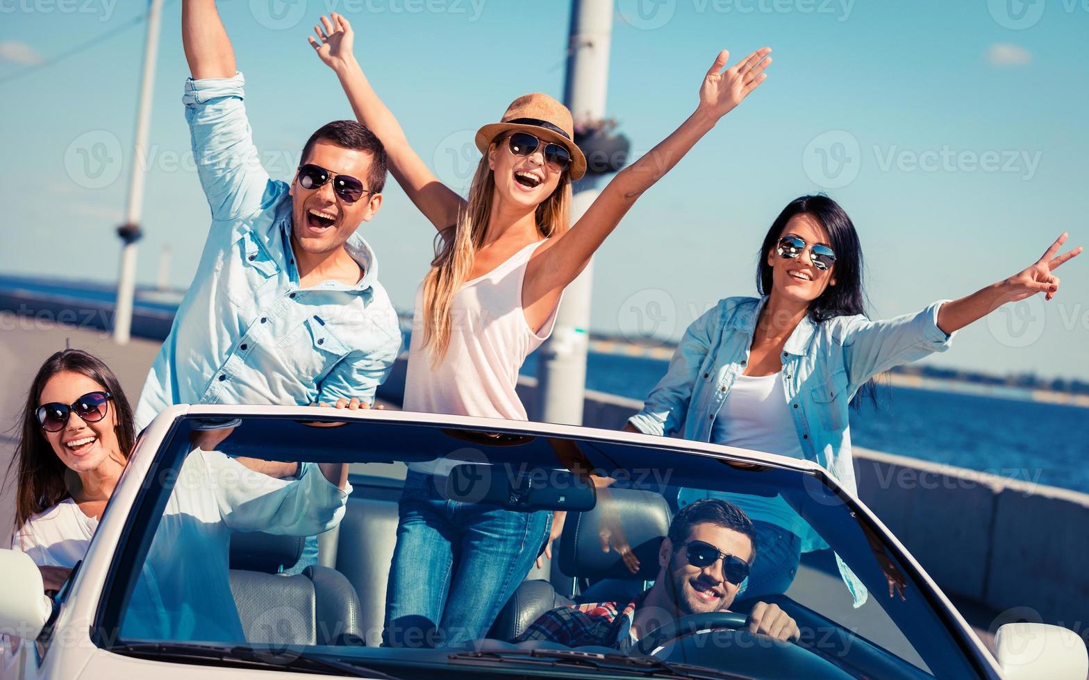 Friends in convertible. Group of young happy people enjoying road trip in their white convertible and raising their arms photo