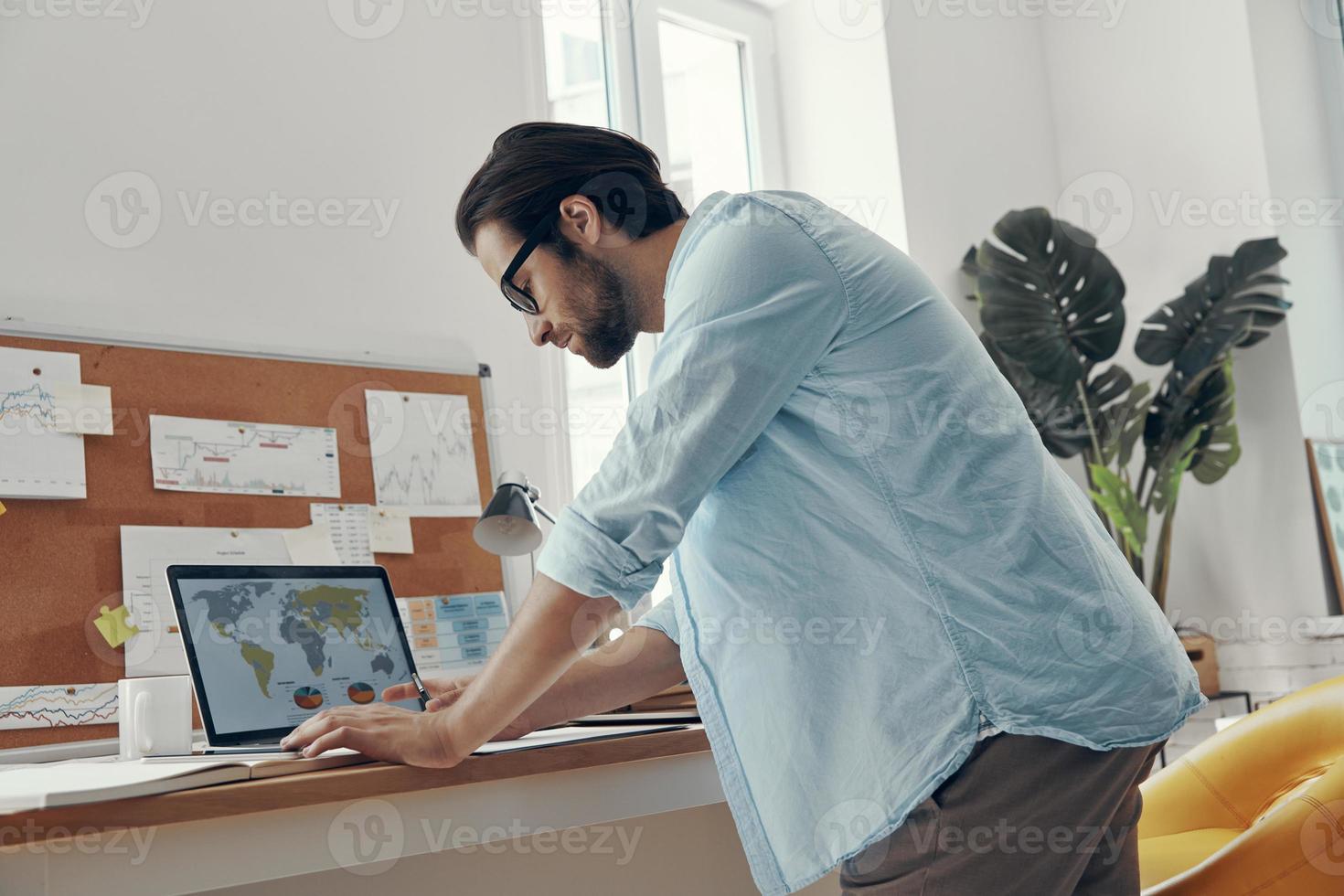 Confident young man using laptop while standing near his working place photo
