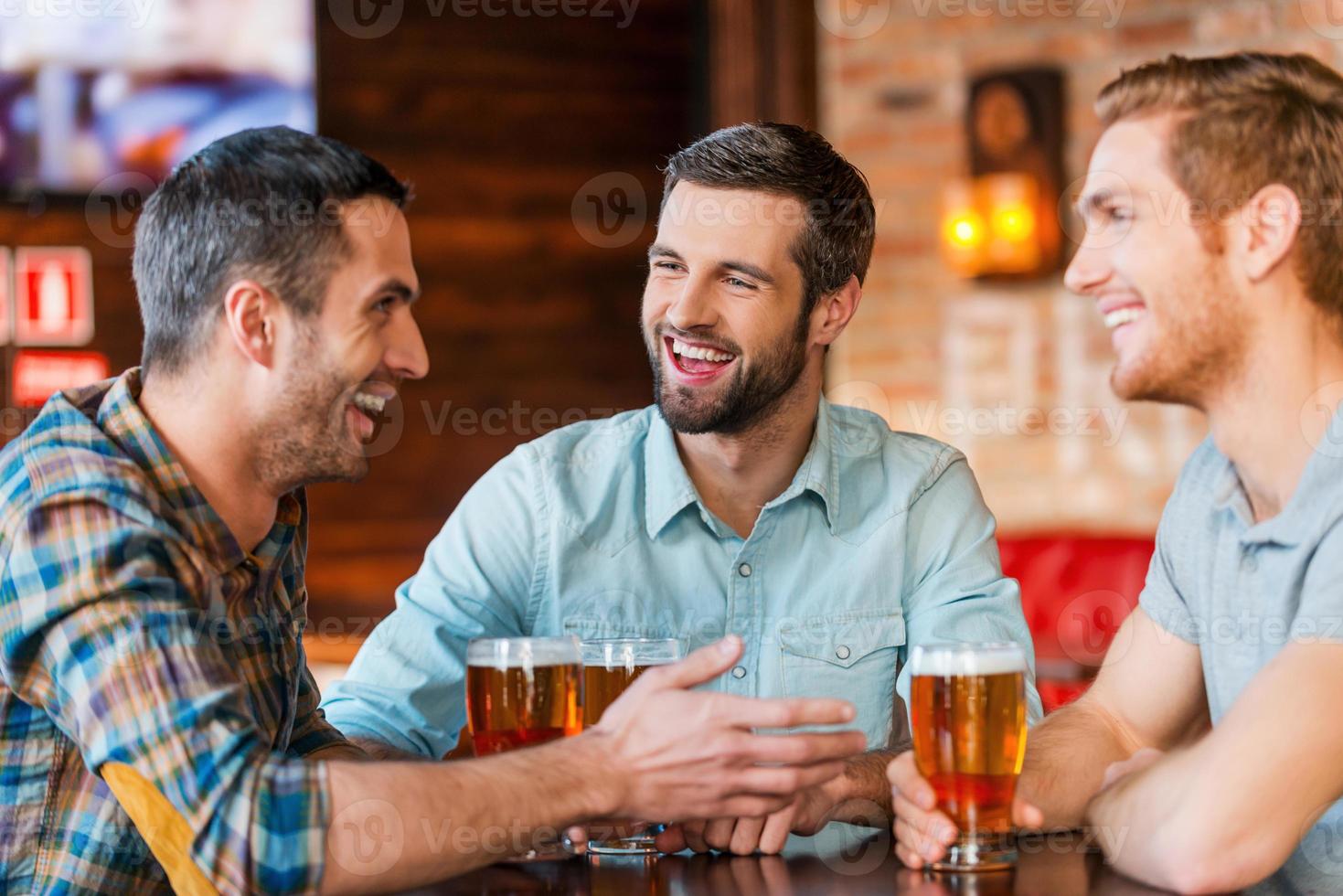 Meeting with the best friends. Three happy young men in casual wear talking and drinking beer while sitting in bar together photo