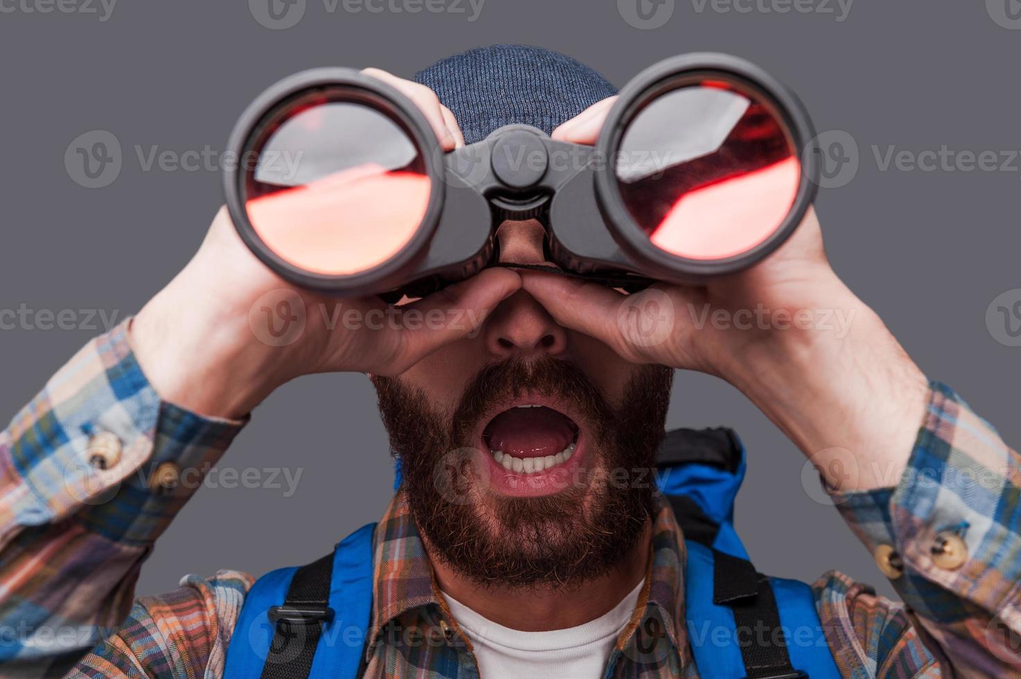 sorprendido joven barbudo llevando mochila y mirando a través de binoculares mientras está de pie contra el fondo gris foto