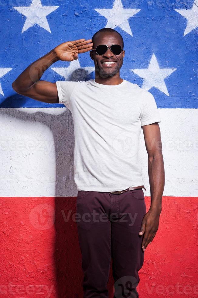 Yes sir Happy young African man in sunglasses holding hand near forehead and smiling while standing against American flag photo