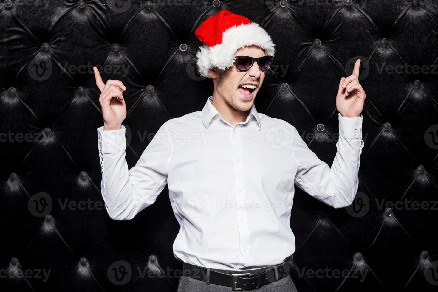 Are you ready to party Handsome young man in sunglasses and Santa hat pointing up and expressing positivity while standing against black background photo