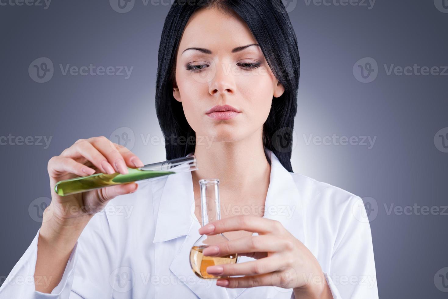 The goal is to find the cure from this disease. Female doctor in white uniform holding flasks while standing against grey background photo