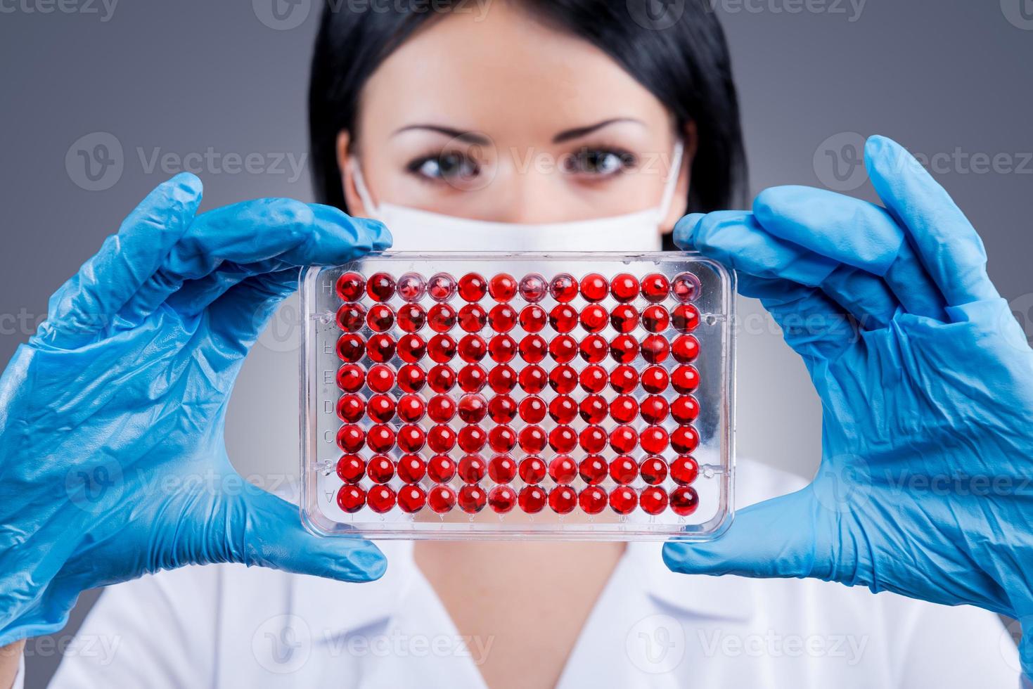 Working in laboratory is what I love. Confident female doctor in white uniform holding micro titer plate and looking at camera while standing against grey background photo