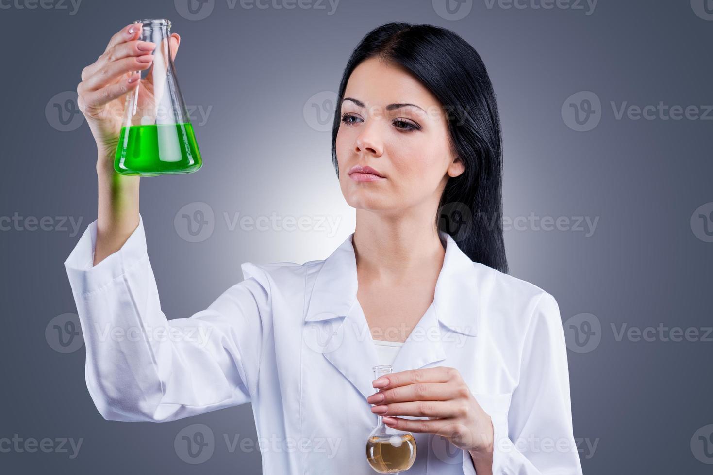 Research studies.  Beautiful  female doctor in white uniform holding flasks while standing against grey background photo