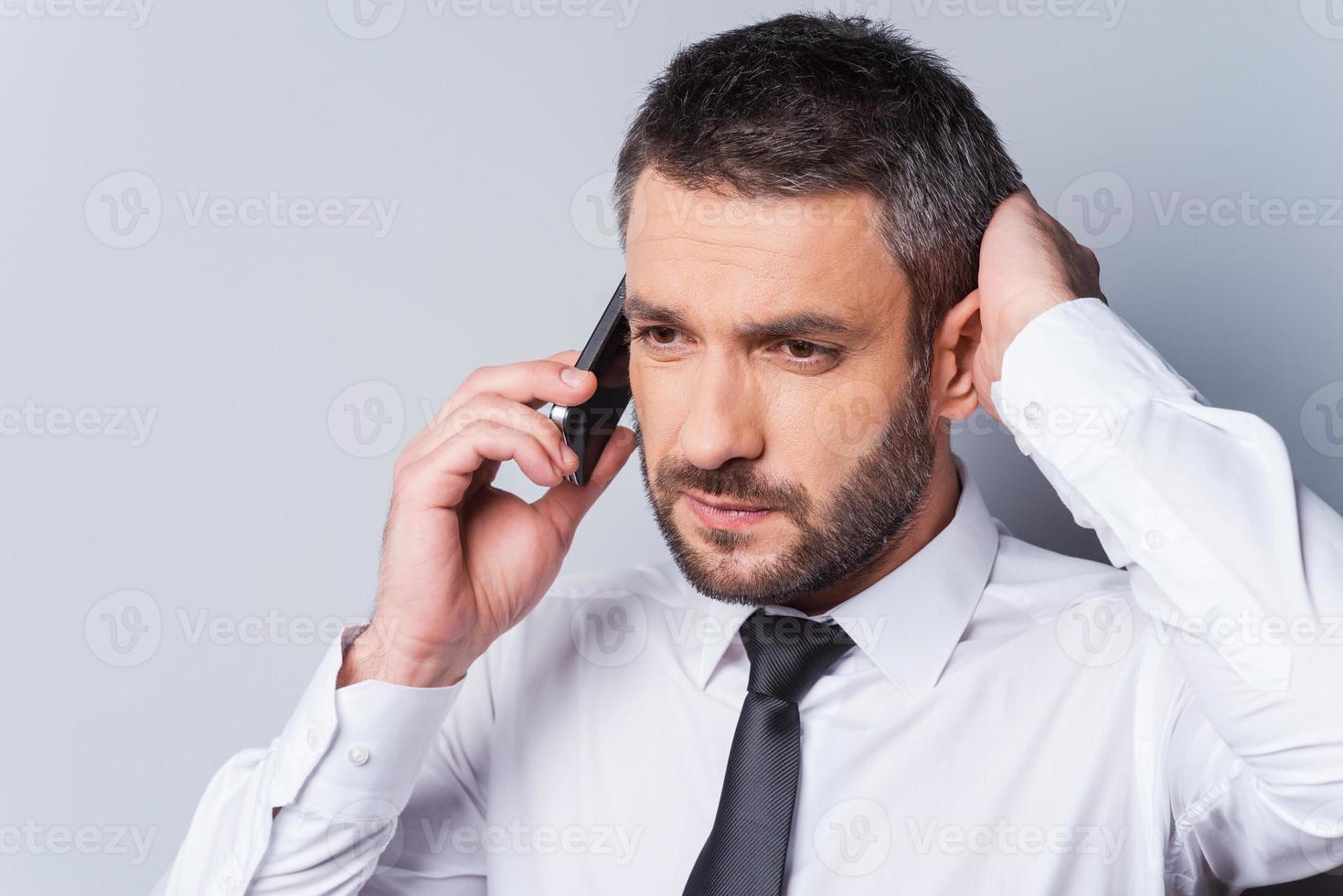 Bad news. Frustrated mature man in shirt and tie talking on the mobile phone and holding hand in hair while standing against grey background photo