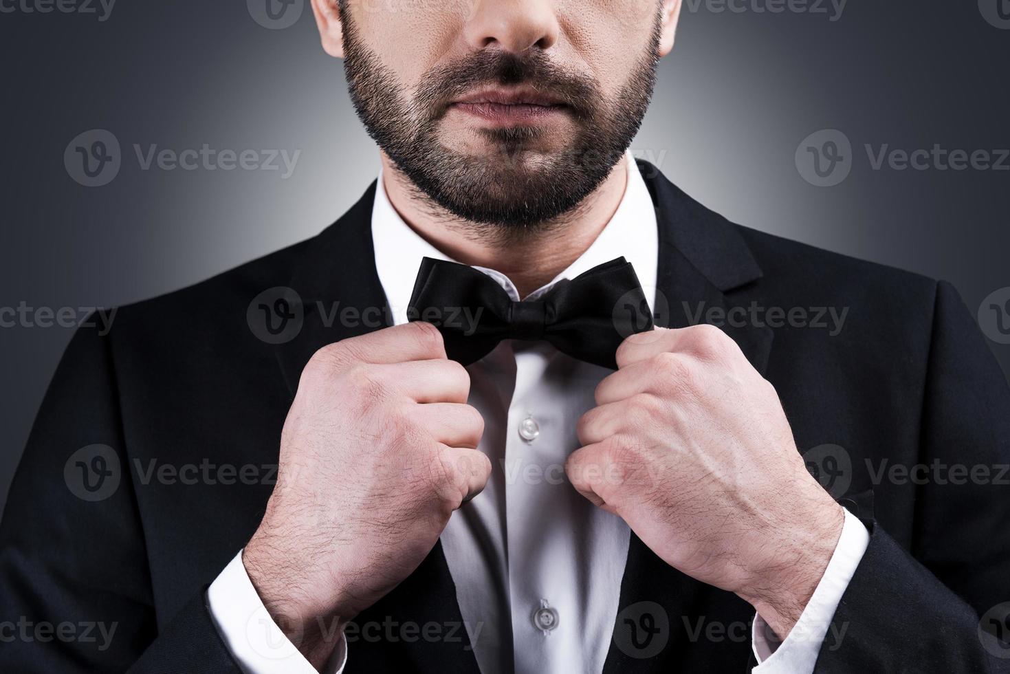 Perfect style. Close-up of handsome mature man in formalwear adjusting his bow tie while standing against grey background photo
