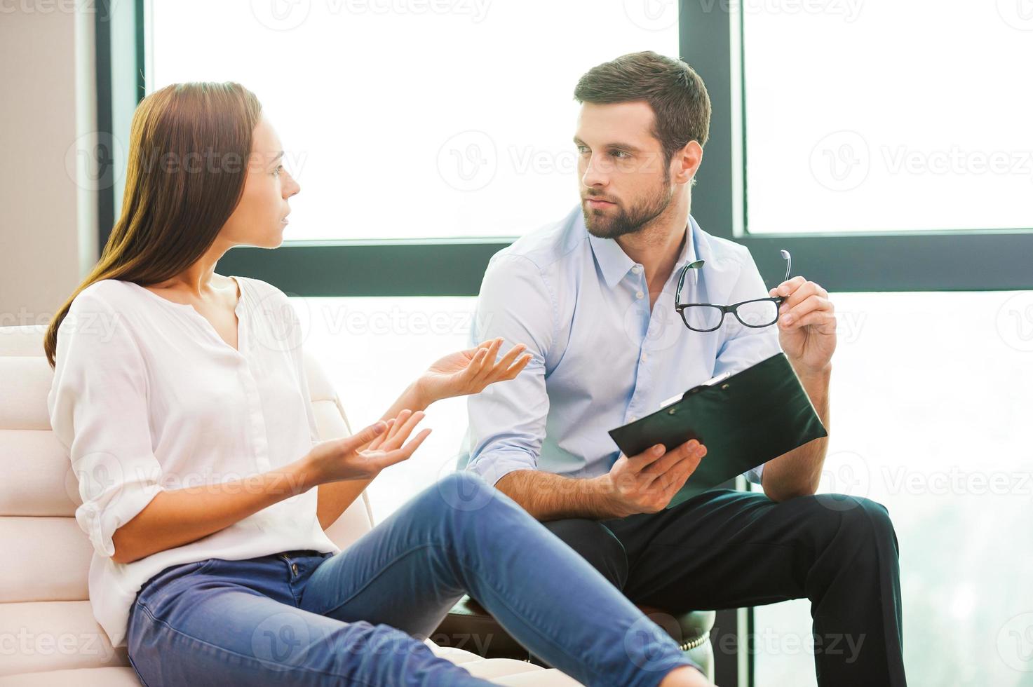 Sharing problems with psychiatrist. Worried young woman sitting at the chair and gesturing while male psychiatrist sitting close to her and holding clipboard photo