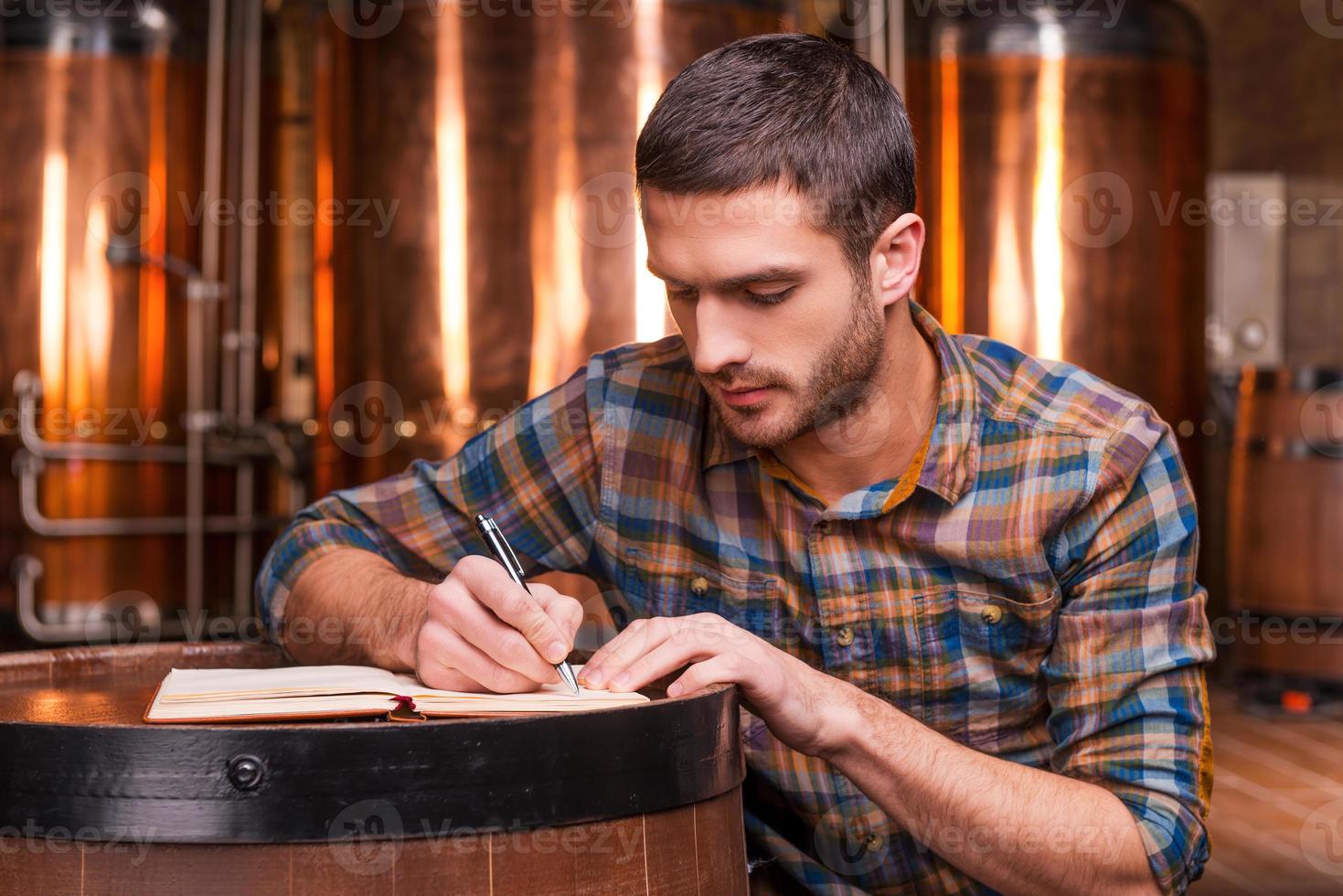 Planning his beer business. Confident young man in casual shirt writing something in his note pad while leaning at the wooden barrel with metal containers in the background photo