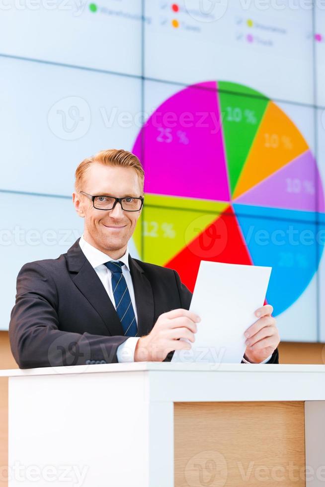 Confident public speaker. Confident young man in formalwear standing at the tribune and smiling while making a presentation with projection screen in the background photo