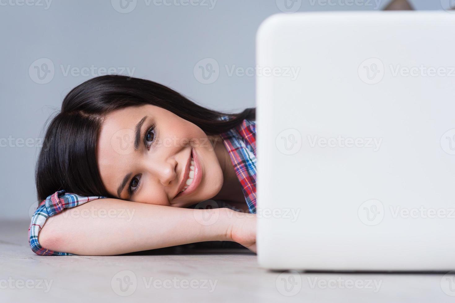 Digital age beauty. Attractive young woman looking out of laptop and smiling while lying on the hardwood floor photo