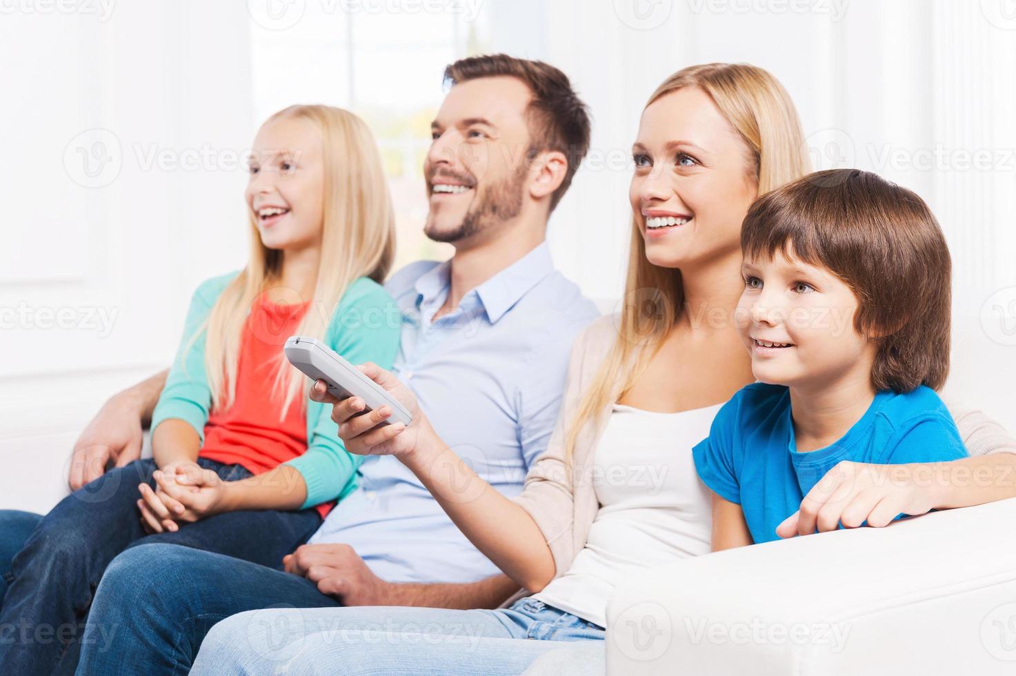 Watching their favorite show. Side View of happy family of four bonding to each other and smiling while watching TV at home together photo
