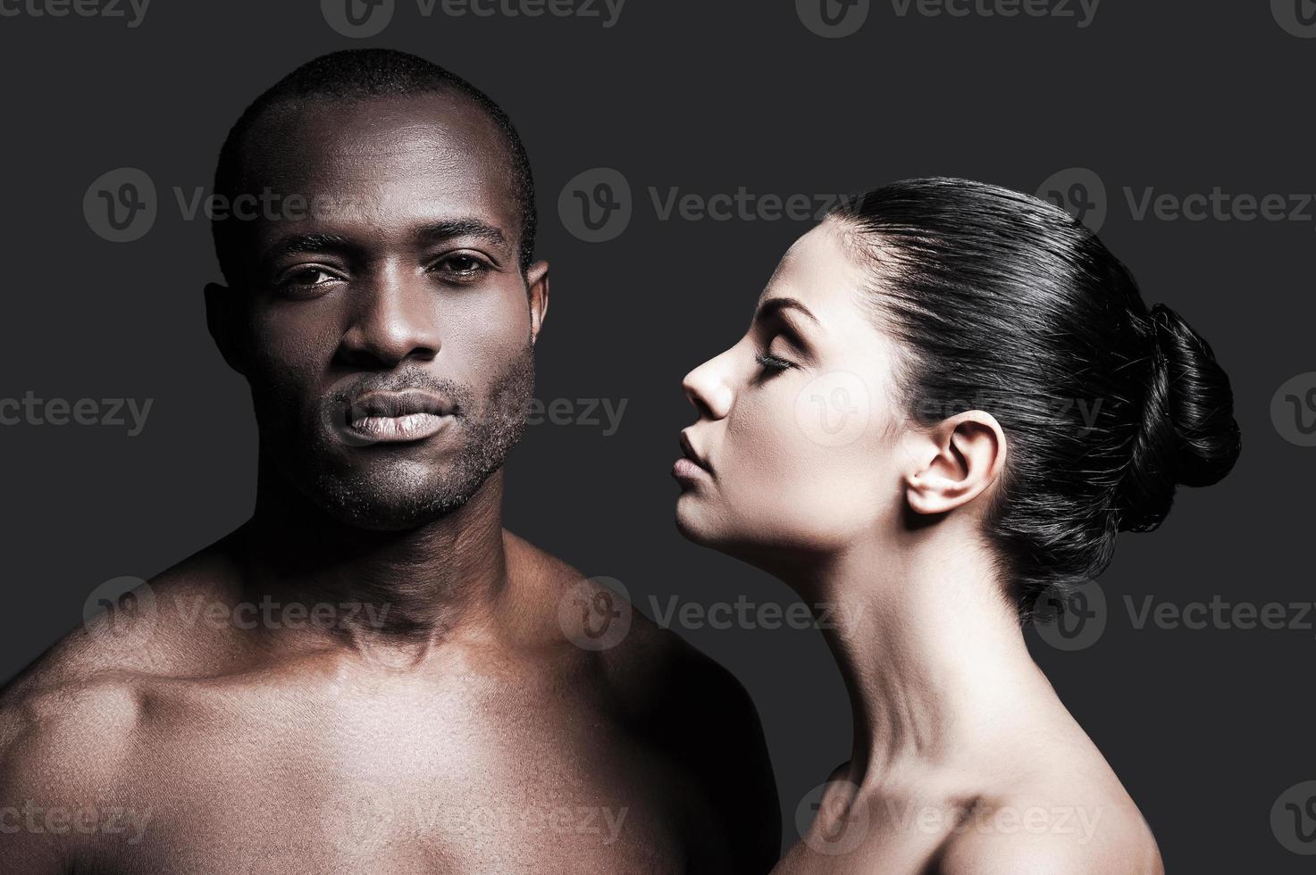 Black and white. Portrait of shirtless African man and Caucasian woman bonding to each other while standing against grey background photo