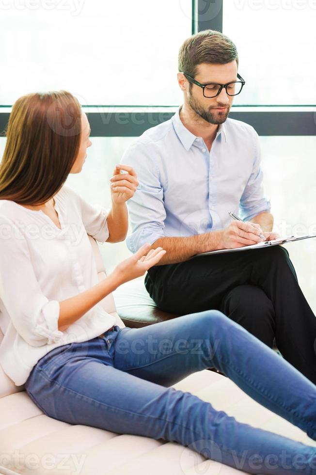 Sharing problems with professional. Worried young woman sitting at the chair and gesturing while male psychiatrist sitting close to her and writing something in his clipboard photo