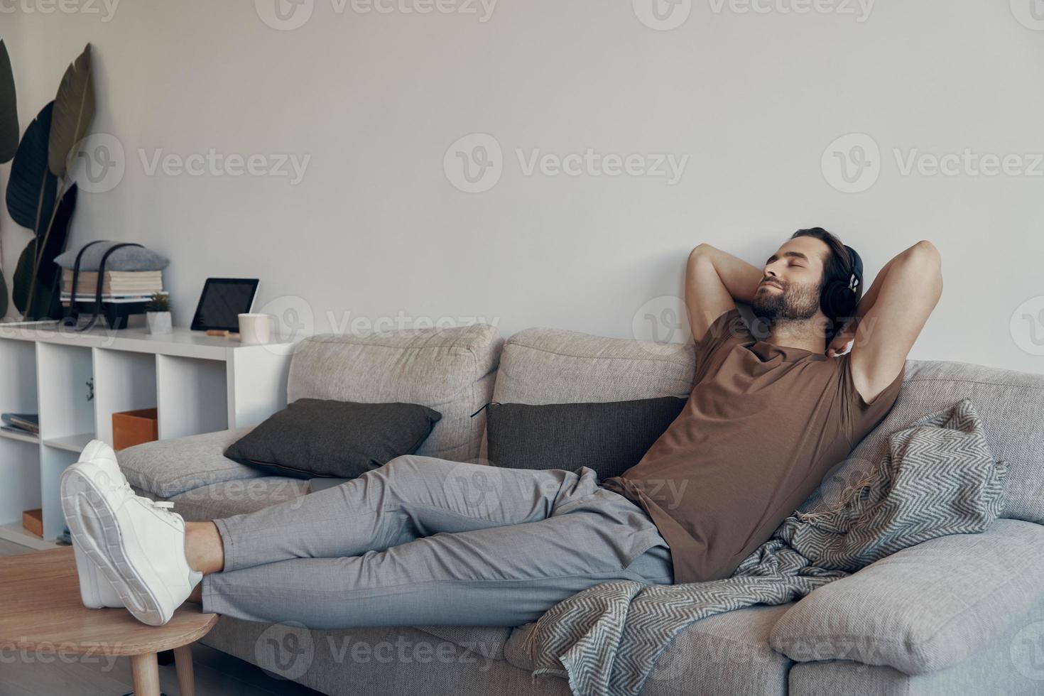 Handsome young man in headphones holding hands behind head while relaxing on the couch at home photo