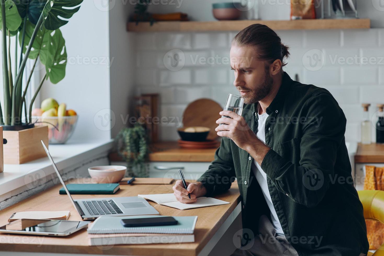 Confident young man making notes and drinking water while working at the domestic kitchen photo