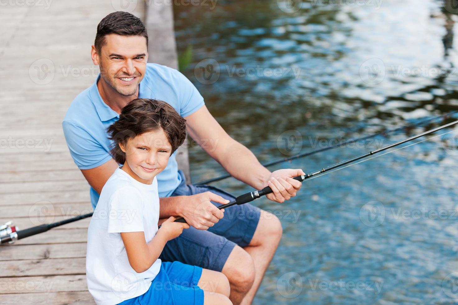 Spending great time together. Top view of father and son looking at camera and smiling while sitting at the quayside with fishing rods laying near them photo