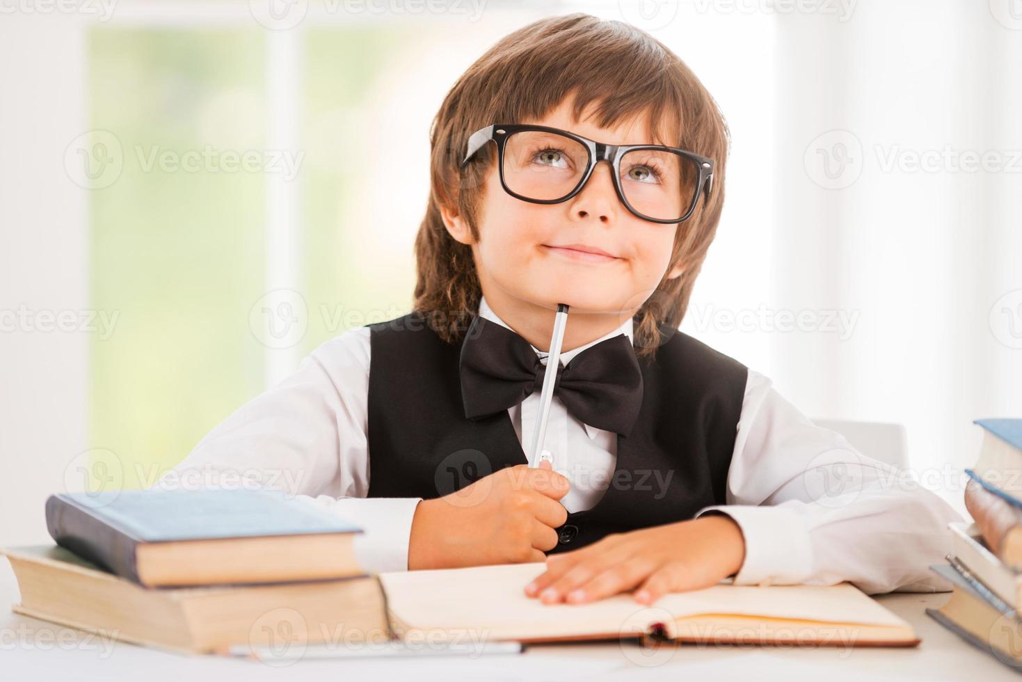 Day dreaming. Little boy leaning his face on pen and looking away while sitting at the desk photo