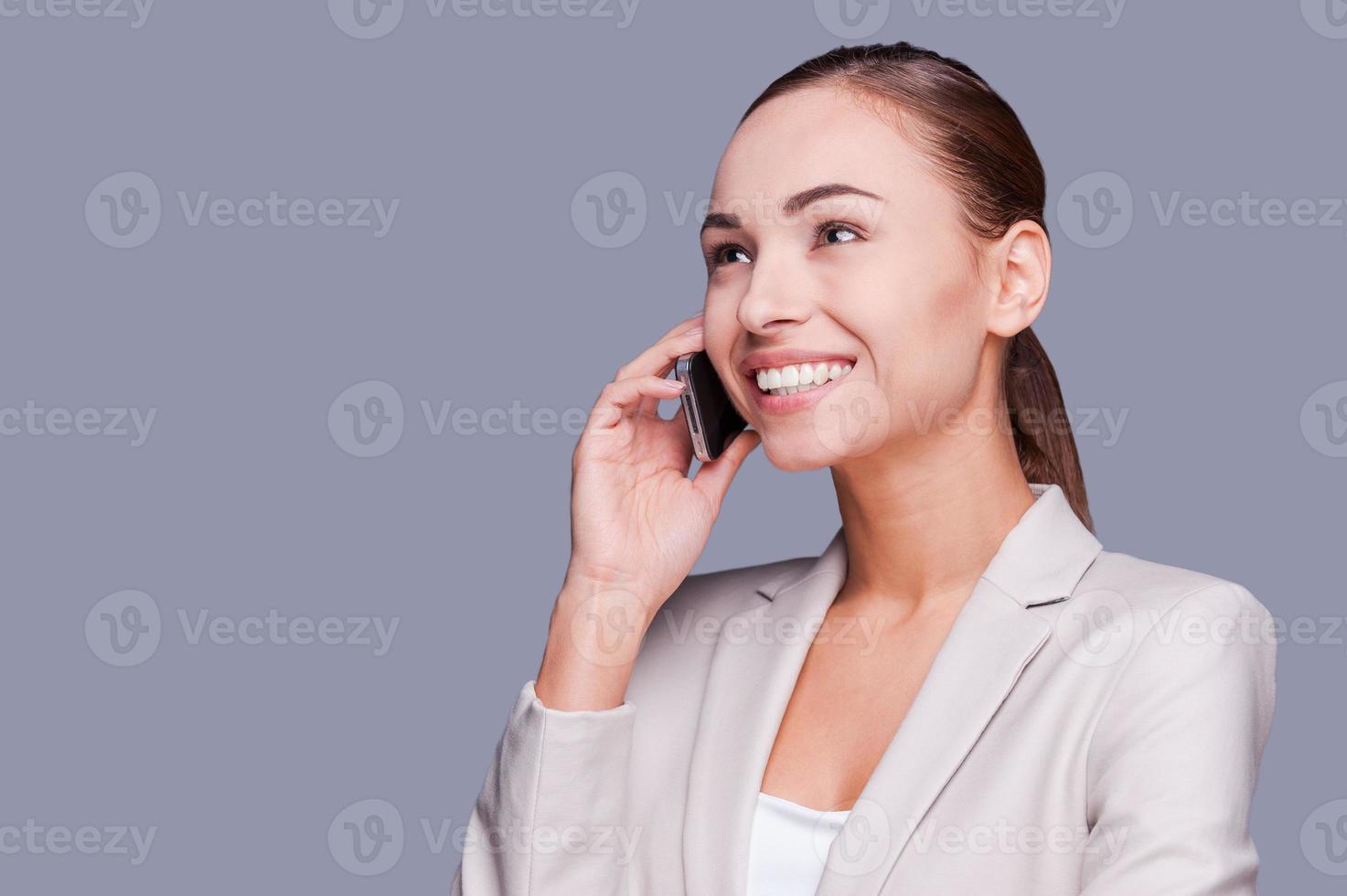 Business talk. Beautiful young businesswoman talking on the mobile phone and smiling while standing against grey background photo