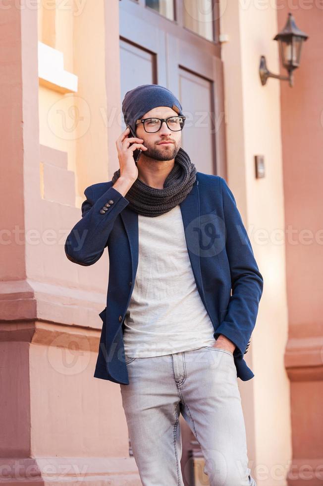 Handsome on the phone. Handsome young man in smart casual wear talking on the mobile phone while standing in front of the house entrance and holding coffee cup photo