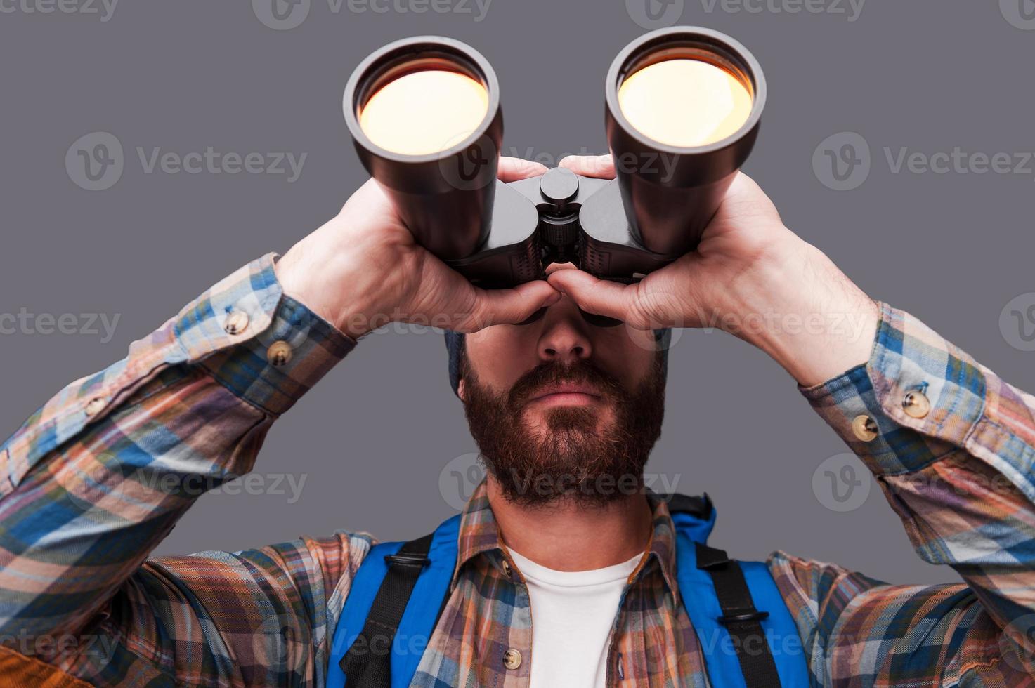Exploring new places. Confident young bearded man carrying backpack and looking through binoculars while standing against grey background photo