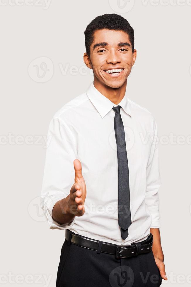 Portrait of cheerful young Afro-American man in formalwear stretching out hand for shaking while standing against grey background photo