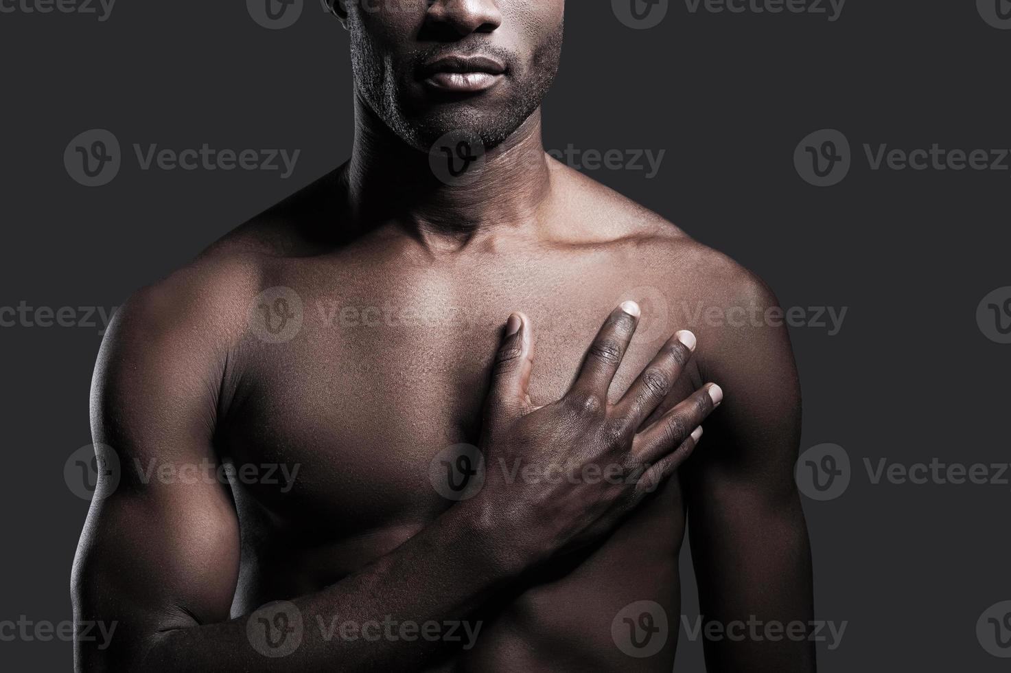It is in my heart. Cropped image of young shirtless African man holding his hand on heart while standing against grey background photo