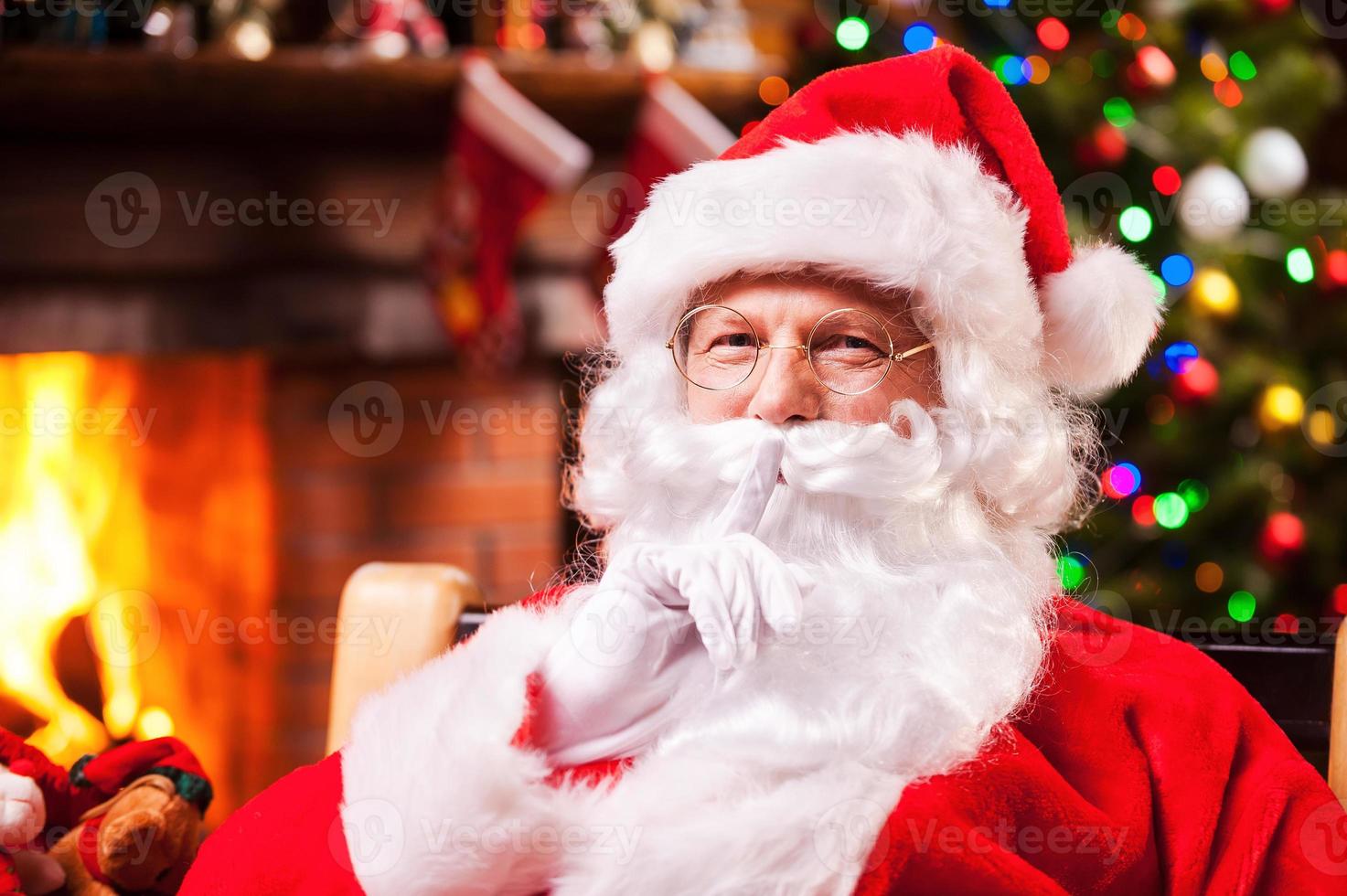 You did not see me Traditional Santa Claus gesturing silence sign while sitting at his chair with fireplace and Christmas Tree in the background photo