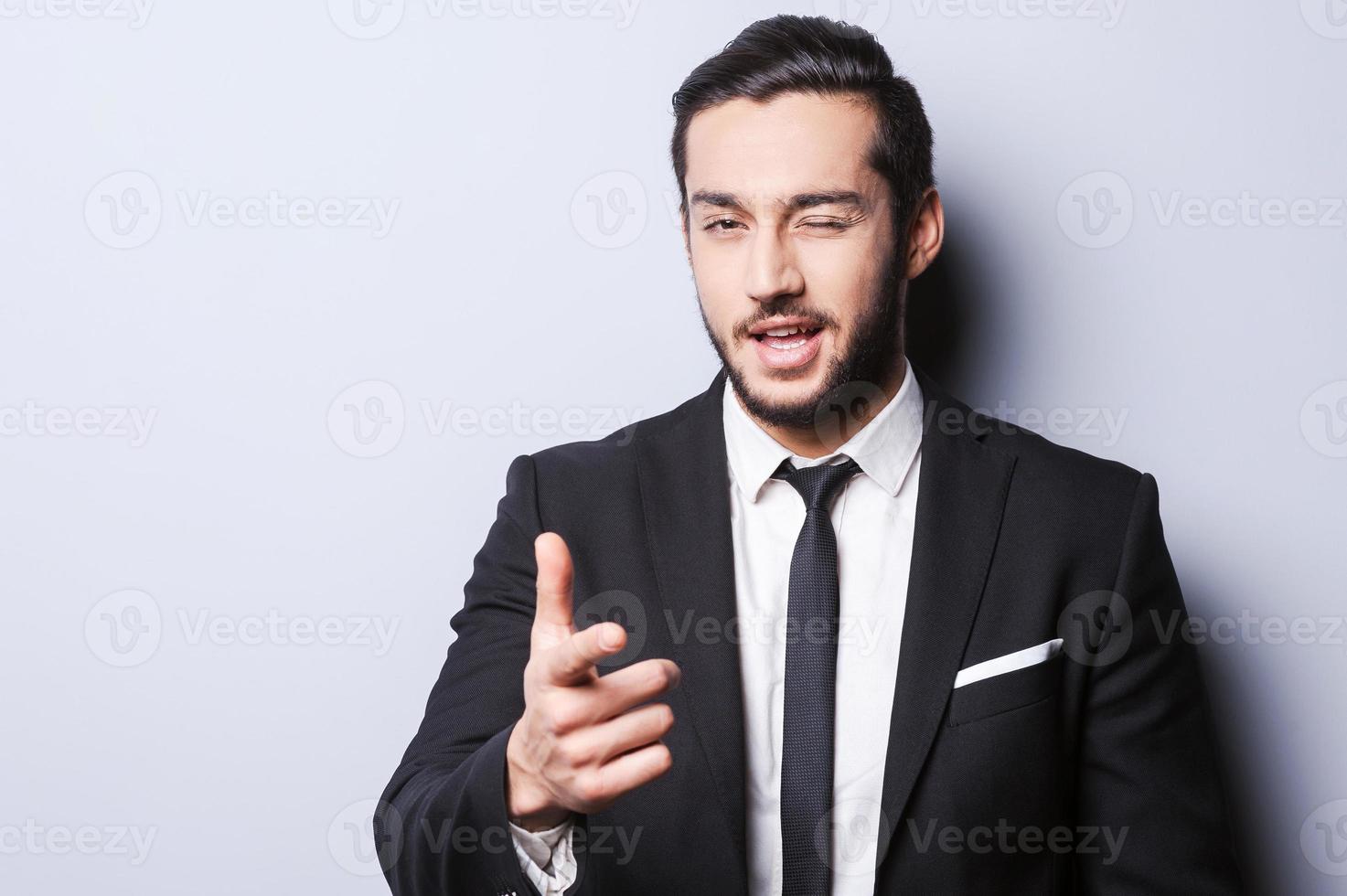 I hope we understand each other. Joyful young man in formalwear winking and pointing on you while standing against grey background photo