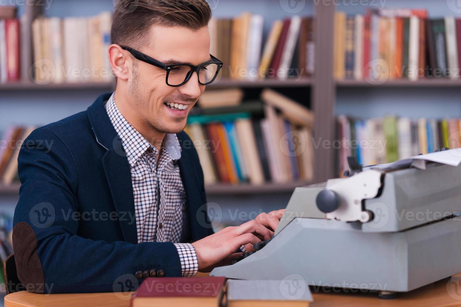 Successful author. Side view of happy young author typing something at the typewriter and smiling while sitting at his working place with bookshelf in the background photo