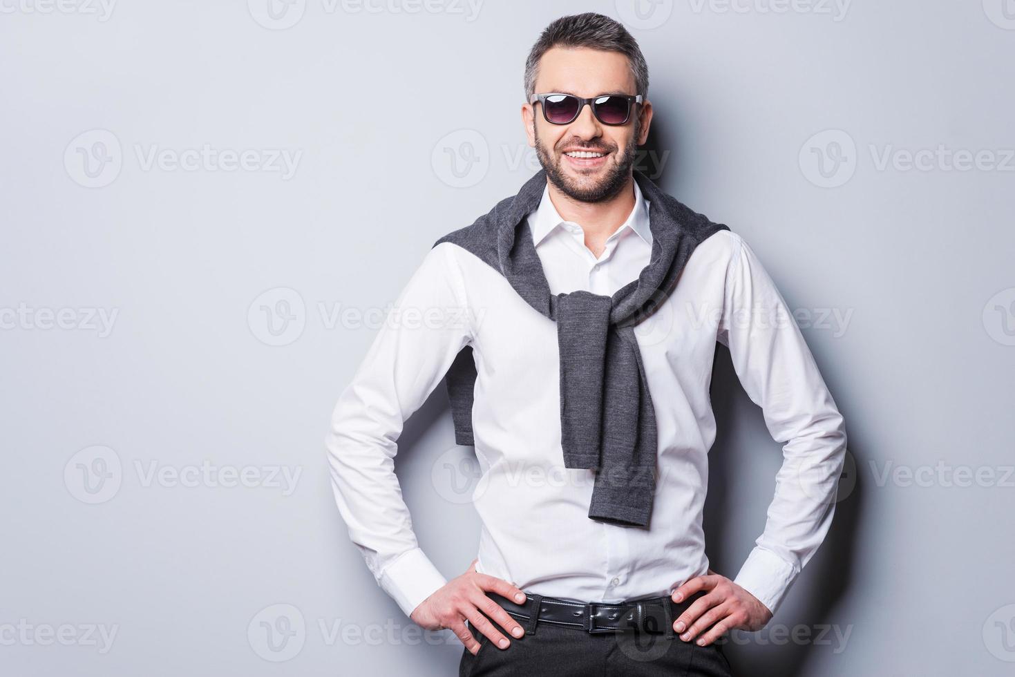 Confident in his perfect style. Handsome young man in smart casual wear and sunglasses looking at camera and smiling while holding hands on hip and standing against grey background photo