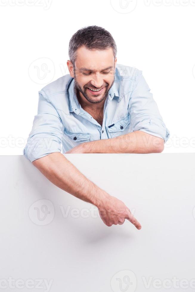 Look at that Happy young man in casual wear leaning at copy space and pointing it while standing against white background photo