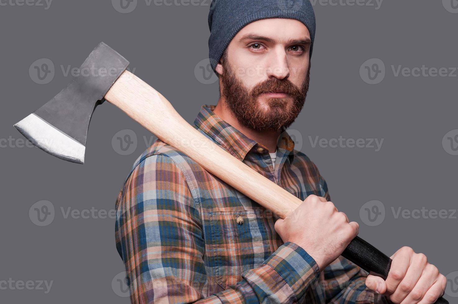 Listo para trabajar. vista lateral de un joven barbudo confiado sosteniendo un hacha grande y mirando la cámara mientras se enfrenta a un fondo gris foto