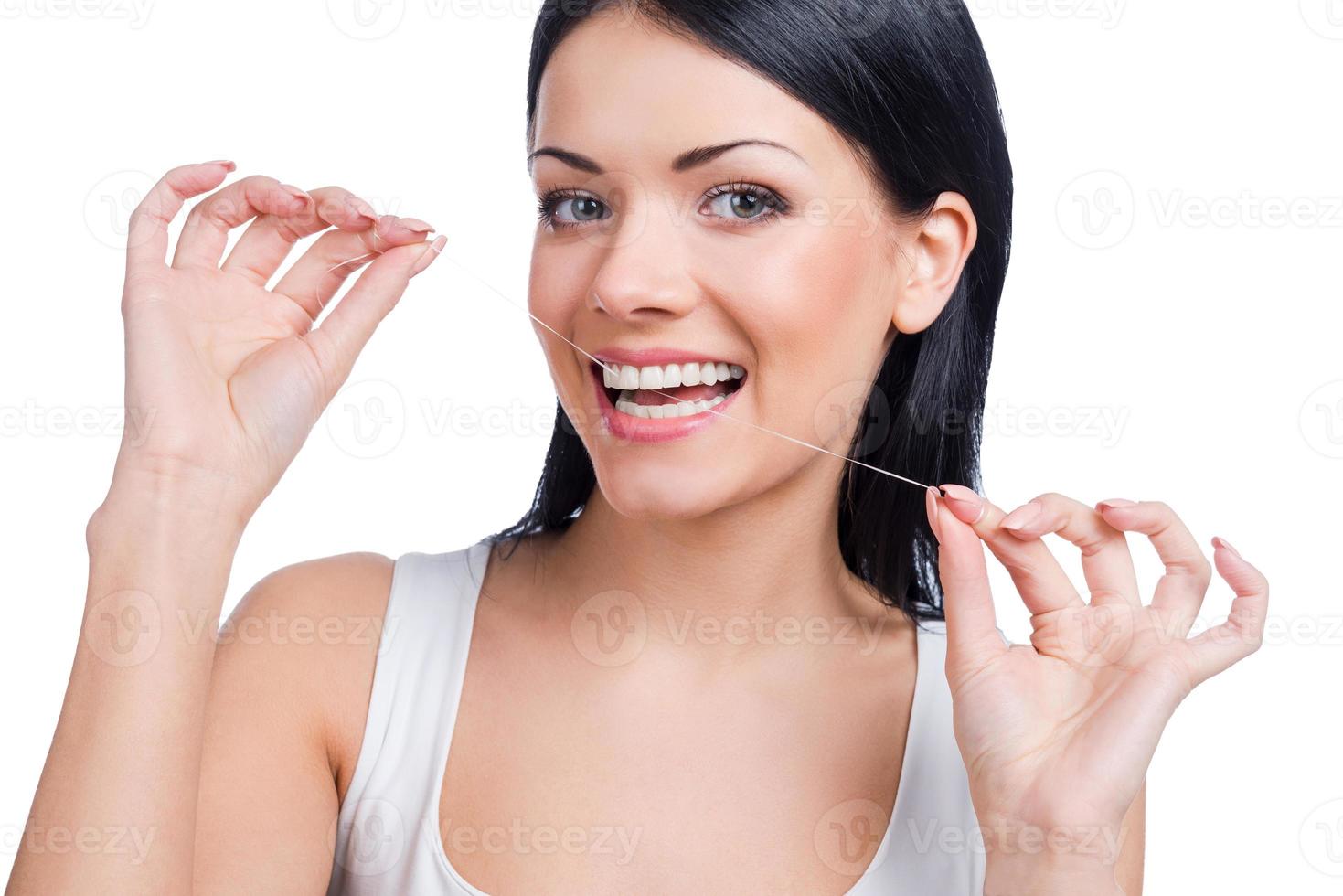 Dental cleaning. Beautiful young woman holding dental floss in her mouth and smiling at camera while standing against white background photo