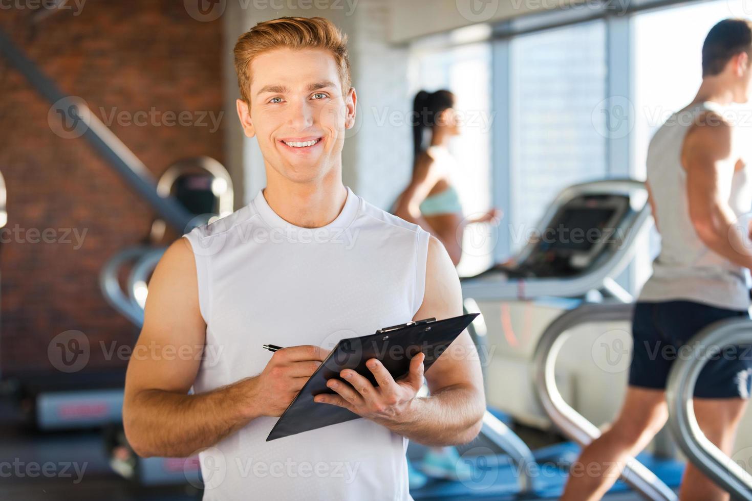 I will make you healthy Handsome young man holding clipboard and smiling while people running on treadmill in the background photo