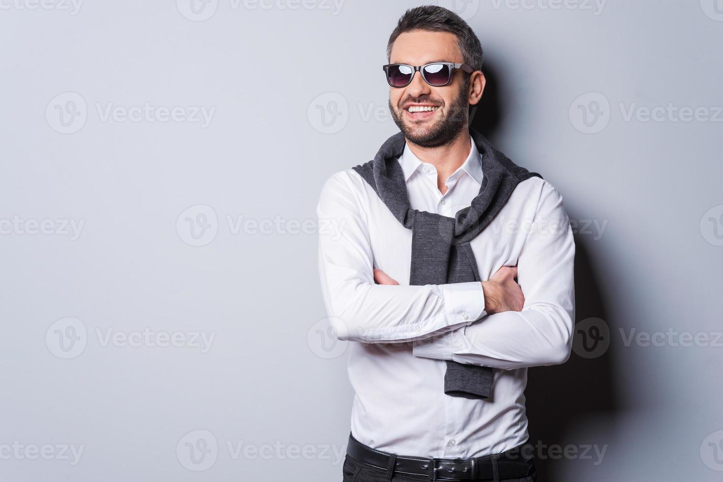 Cheerful handsome. Handsome young man in sunglasses and smart casual wear keeping arms crossed and smiling while standing against grey background photo