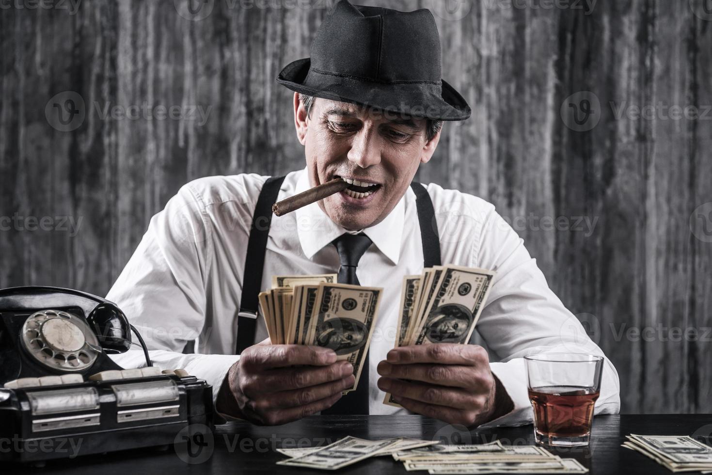 Money and power. Senior gangster in shirt and suspenders counting money and smiling while sitting at the table photo