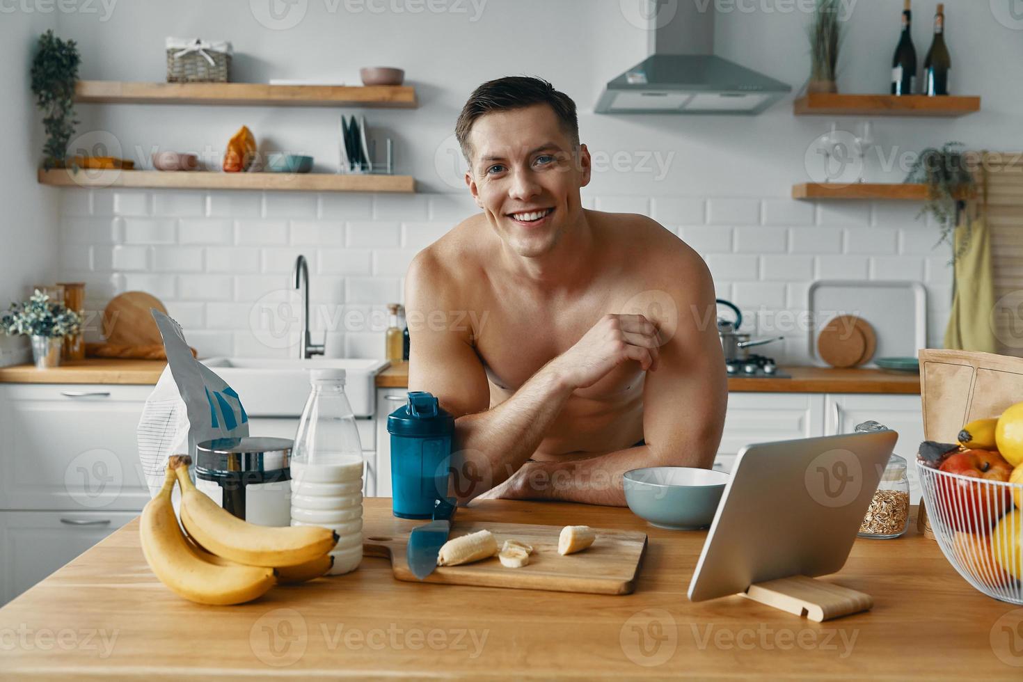 Confident young man looking at camera and smiling while preparing healthy food at the kitchen photo
