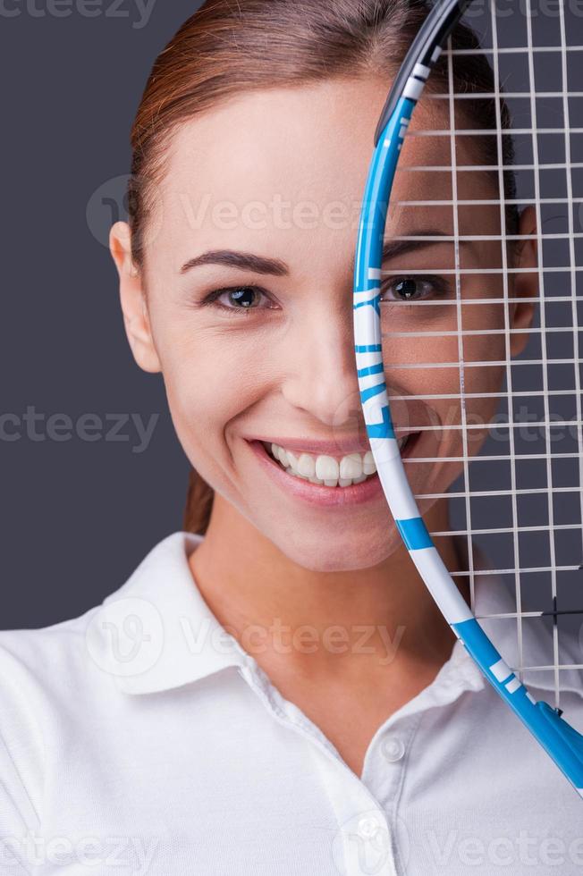 Pretty competitive.  Beautiful young women in sports clothes holding tennis racket in front of half of her face and looking at camera while standing against grey background photo