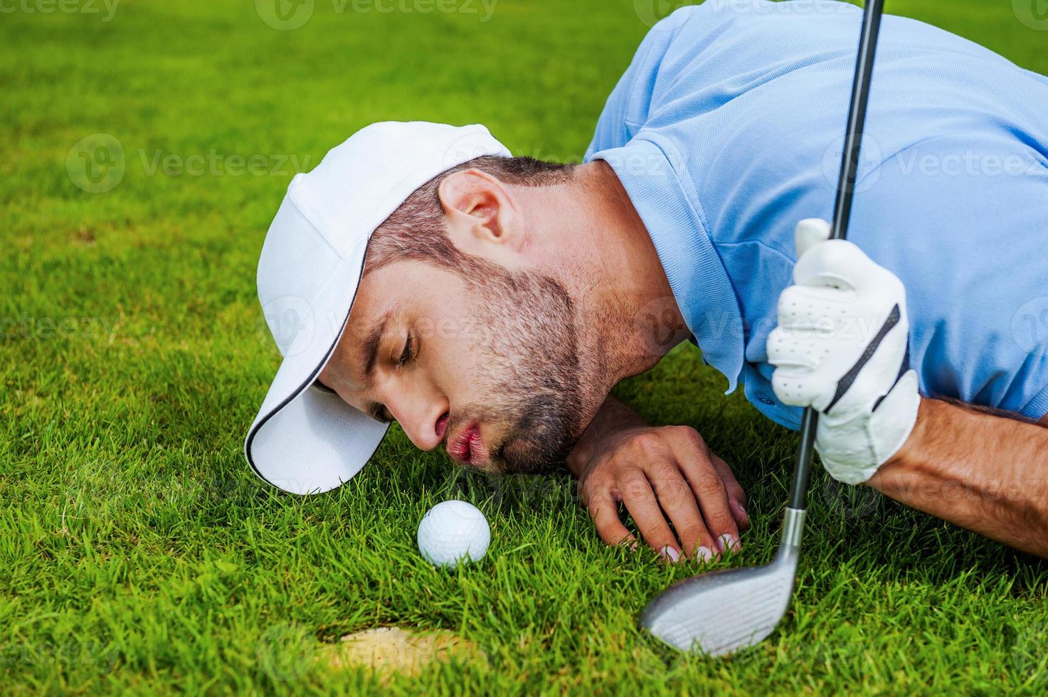 Golf cheats. Close-up of young golfer blowing at the ball while lying near the hole photo