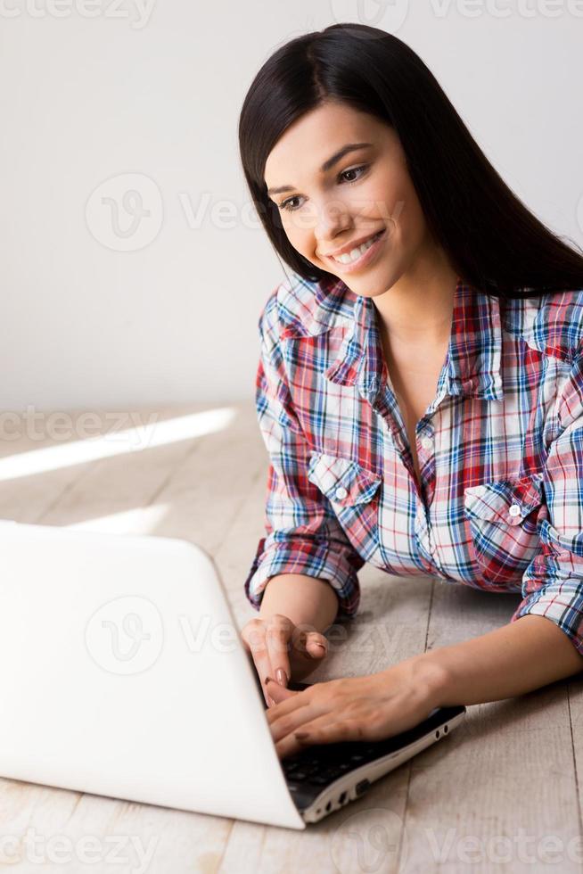 Spending time in the net. Beautiful young woman working on laptop and smiling while lying on the hardwood floor photo
