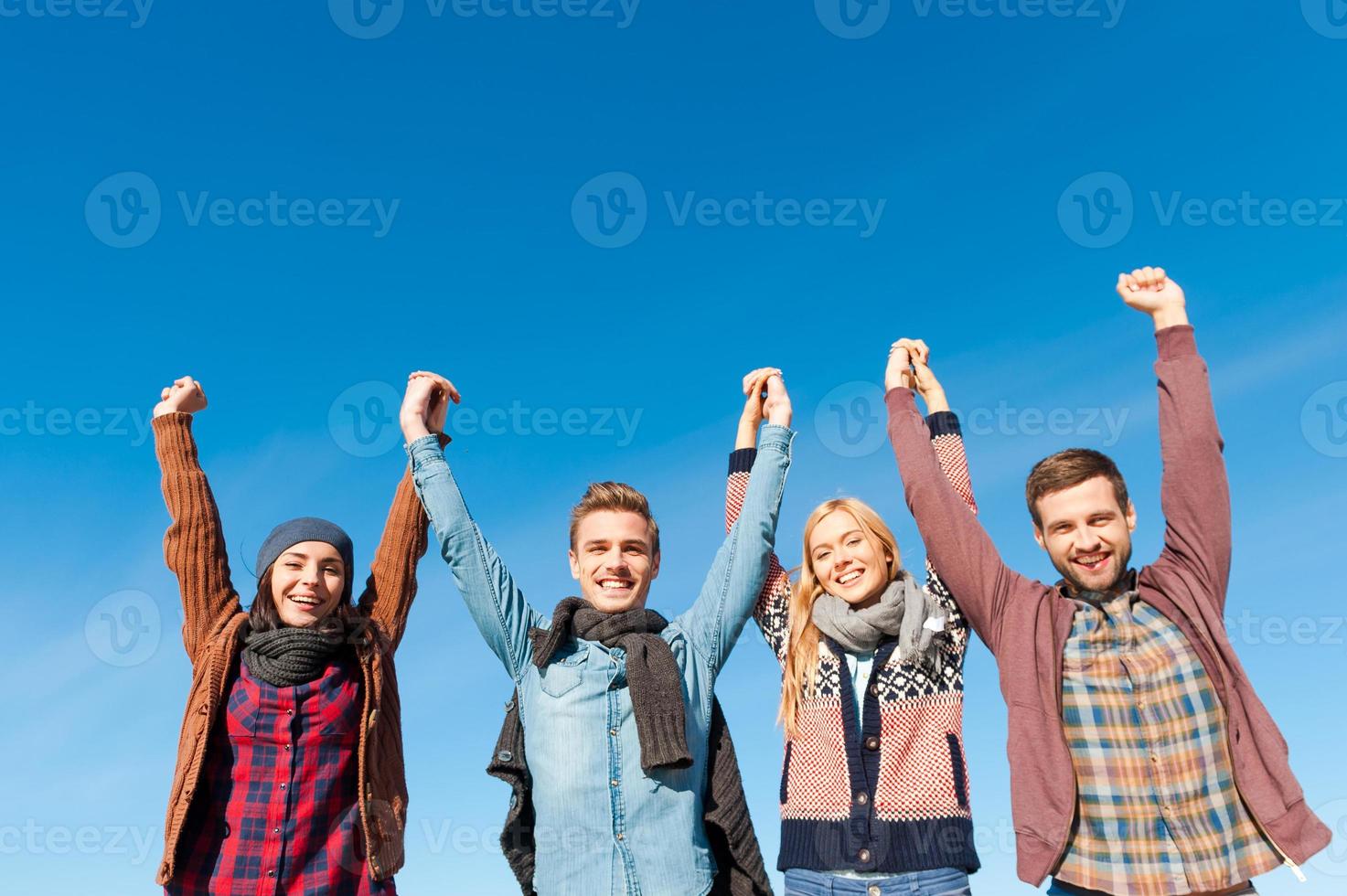 Forever young. Low angle view of four young happy people holding hands and raising them up with blue sky as background photo