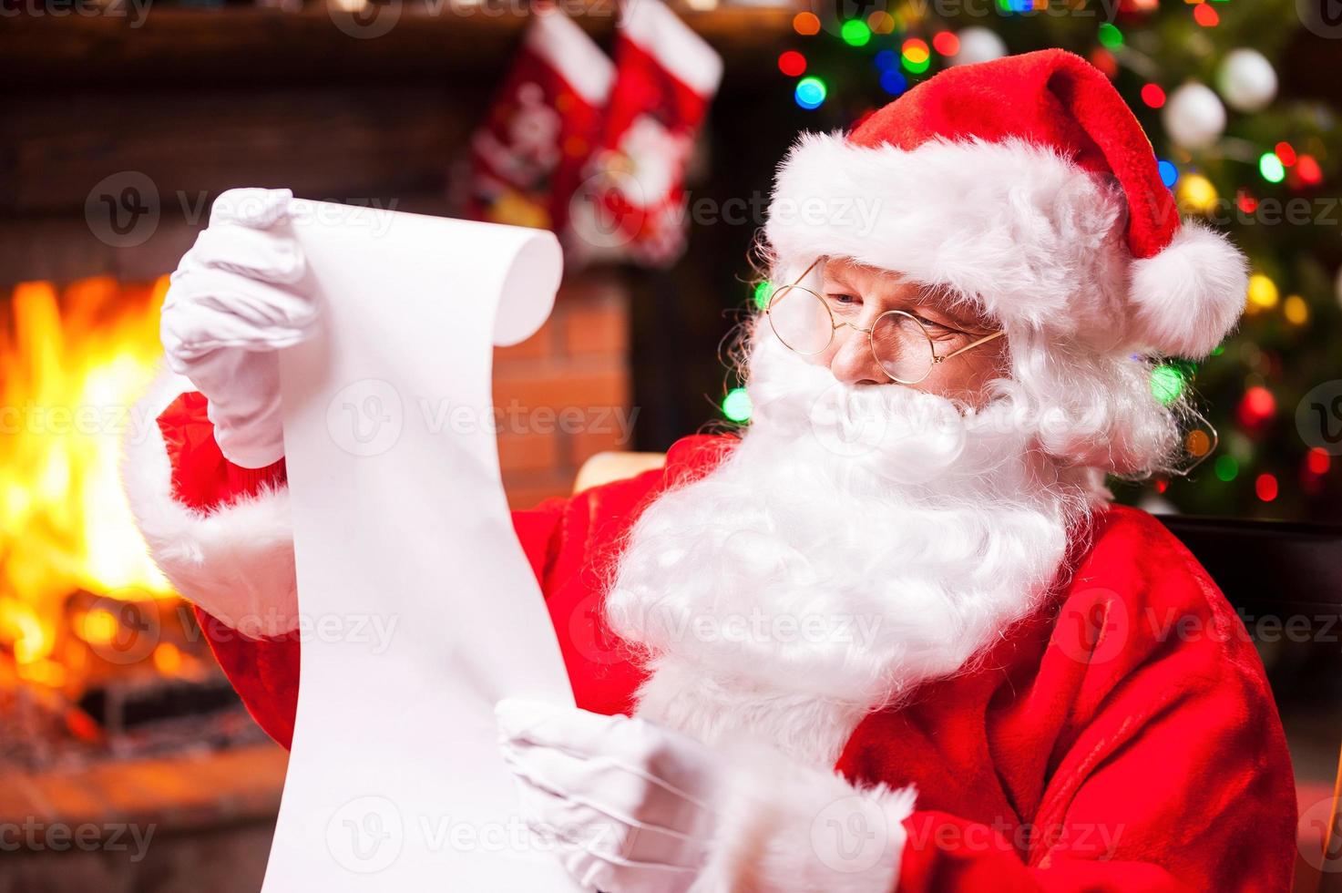 quien fue travieso este año tradicional santa claus sentado en su silla y leyendo una carta con chimenea y árbol de navidad en el fondo foto