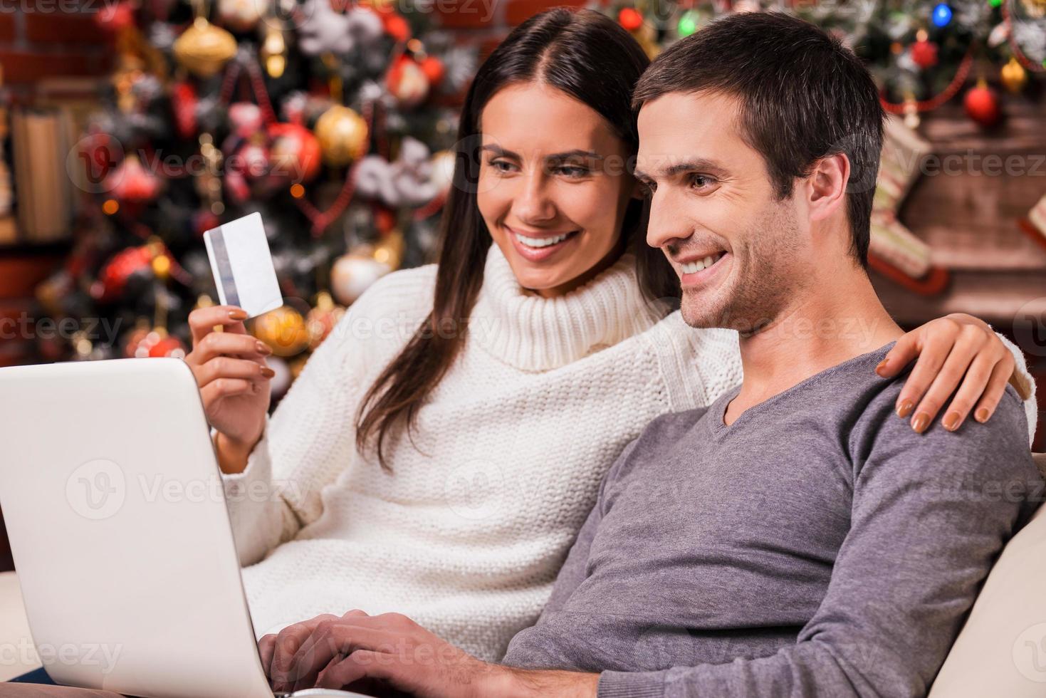 Taking advantages of Christmas sales. Beautiful young couple buying online while using computer together with Christmas Tree in the background photo